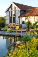 Vue sur l'étang de baignade à la terrasse en bois, à l'extérieur de la salle à manger avec table et chaises et grange convertie avec la chaleur du soleil d'été reflétée sur le bâtiment - Carpe Diem, Bressingham, Norfolk