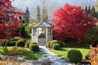 Un chemin marqué et un pavillon en bois peint en blanc à côté de Acer palmatum - érables japonais aux couleurs de l'automne. Buxus - Boîtes de balles