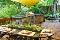 Cuisine extérieure d'été avec grill et évier sur terrasse en bois. Chaises et table en fer forgé avec centre de table en guirlande succulente.