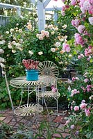 Coin salon sous pergola avec rosiers grimpants, table et chaises de jardin en métal, Rosa 'Cornelia', Rosa 'Buff Beauty', Rosa 'Ghislaine de Féligonde', sedum en pot bleu, sol en brique