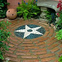 Cour circulaire avec banc en pierre et fougères entourant le sol carrelé avec motif central en étoile créé à partir de galets blancs et gris.