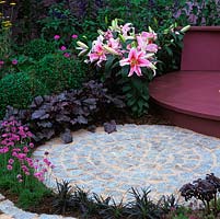 Patios imbriqués de terrasse en bois de couleur grenat circulaire bordée de lys Stargazer, de heuchera, de balles en boîte, d'Ophiopogon planiscapus Nigrescens et de roses de mer.