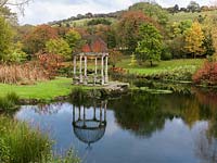 Rotonde de pierre à côté d'un lac calme bordé de feuilles rouges de Darmera peltata, de joncs, d'iris, de saules, de bambous, d'aulnes et de Liquidambar styraciflua. Les collines du Dorset se dressent derrière.