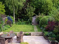 Un jardin familial avec terrasse en pierre avec coin salon et salle à manger. Deux marches mènent à la pelouse. Vu de la fenêtre à l'étage. Meubles en osier