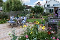 Terrasse en bois avec barbecue, table et chaises, bordée de pots et de parterres de fleurs.