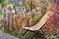 Chaise de terrasse en bois parmi les vivaces et les graminées en automne: Pennisetum, Crocosmia 'Lucifer', Aster, Sedum, Panicum virgatum.