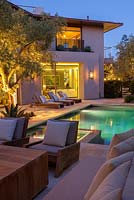 Vue sur la piscine moderne et la maison avec des chaises longues et un olivier mature au crépuscule.