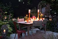 La table est préparée pour le barbecue d'été avec un vase de roses et des bougies. Des bougeoirs en verre dans des parterres illuminent la plantation de gauche à droite, hosta, epimedium, Hydrangea 'Annabelle', lysimachia, Verbena bonariensis et Geranium 'Rozanne '. Reflets dans le miroir mural de la porte de la cour.