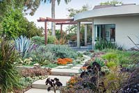 Vue sur un chemin en béton et des parterres de fleurs mixtes vers une maison moderne et un coin repas extérieur. Encinitas, Californie, États-Unis. Août.