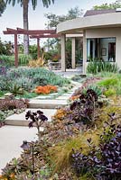 Vue sur un chemin en béton et des parterres de fleurs mixtes vers une maison moderne et un coin repas extérieur. Jardin de Debora Carl, Encinitas, Californie, USA. Août.