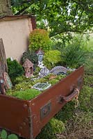 Une scène de jardin miniature dans une valise vintage faite de mousse, de cailloux décoratifs, de coquillages, de figurines d'animaux et de structures, d'écorce d'arbre, de petits conifères et d'éclairage LED