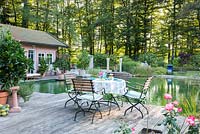 Vue depuis la terrasse en bois avec des meubles de jardin en bois et en fer sur la piscine naturelle jusqu'aux bains. En arrière-plan, une petite zone boisée