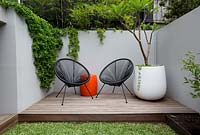 Une terrasse en bois avec deux chaises Acapulco noires, une table à tambour en plastique orange, un pot rond en plastique blanc avec un Plumeria, des frangipaniers devant un mur de soutènement en ciment peint en gris. Avec des plantes succulentes débordant sur le mur et un écran de tisserands élancés en bambou.