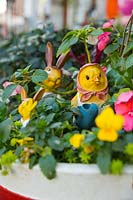 Un pot avec des altos jaunes, des pensées, des Impatiens roses, deux lapins et un poussin avec des visages jaunes et des décorations dans le pot.