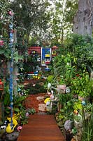 Vue depuis la partie inférieure du jardin jusqu'à la clôture arrière, avec un platelage en bois de forme irrégulière, une collection éclectique de pots, des ornements de jardin, des jouets et des plantes aimant l'ombre.