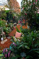Vue depuis une section surélevée du jardin donnant sur une terrasse en bois de forme irrégulière et un mélange éclectique de pots, d'ornements de jardin, de plantes aimant l'ombre et d'un siège de jardin à lattes avec un coussin à motifs orange.