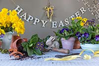 Arrangement de Pâques avec des fleurs de printemps, des outils, des pots, un lapin et un signe de Joyeuses Pâques