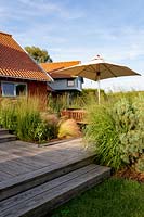 Jardin de prairie à Aldeburgh, Suffolk. Terrasse en bois avec table en bois avec parasol et chaises. La plantation de plates-bandes herbacées comprend: Panicum virgatum - métaux lourds, Molinia caerulea subsp. arundinacea - Transparent, Stipa tenuissima.