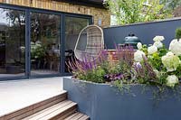 Jardin contemporain de Londres avec vue sur la maison - barbecue sur mesure sur terrasse avec parterre de fleurs surélevé gris. La plantation comprend le smoothie aux baies de Heuchera, la Salvia caradonna, l'Hydrangea anabelle, le Geranium johnsons blue.