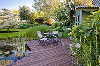 Espace aménagé près d'un étang avec table et chaises, vue sur les parterres de fleurs, la pelouse et les spécimens d'arbres