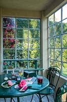 Une serre froide en fer et en verre, maintenant utilisée comme véranda avec table et chaises. Fleurs de camélia et nuancier et pots de conserves, vue sur le jardin.