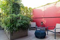 Jardin sur le toit avec une cloison rouge avec des sièges à l'avant, une surface en bois avec le même matériau utilisé pour une grande jardinière d'arbustes qui divise l'espace
