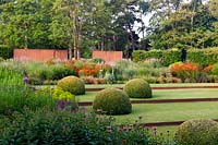 Jardin de campagne moderne: pelouse divisée par une bordure en métal rouillé avec des dômes topiaires, des parterres de fleurs profondes, des murs en métal et des arbres
