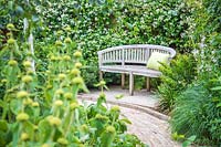 Vue le long du pavage en brique vers banc en bois rustique avec coussin vert en face de clôture recouverte de Trachelospermum jasminoides - Star jasmine