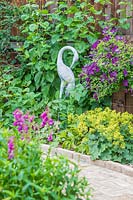 Vue sur parterre de fleurs vers statue de cigogne et parterre de fleurs avec Alchemilla mollis et floraison Clematis viticella sur clôture