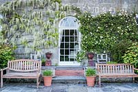 Une porte encadrée de glycines blanches grimpantes et une rose avec des bancs, des urnes et des roses