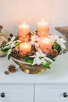 Un arrangement dans un bol doré, avec quatre bougies pilier corail, des fleurs de poinsettia et des cônes