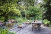 Salle à manger simple en terrasse dans un jardin boisé entouré d'arbres et d'arbustes denses en juin. La plantation comprend des fougères, des bambous et des graminées.