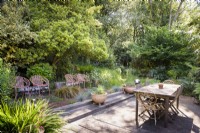 Salle à manger simple avec terrasse entourée d'arbres et d'arbustes denses dans un jardin de cottage en juin. Quatre sièges contemporains en métal orange sont disposés en rangée sur un côté avec des plantations comprenant Carex testacea et bleu festuca.