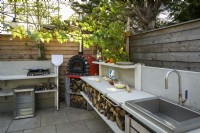 Cuisine extérieure avec four à pizza rouge, évier en métal, plaque de cuisson et rangement en bois dans un petit jardin familial moderne