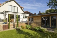 Vue vers maison avec coin salon sur terrasse en bois surélevée et dépendance dans petit jardin familial moderne