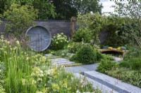 Un endroit pour se retrouver. Hampton Court Flower Festival 2021. Une cour urbaine est plantée dans des tons reposants de gris, vert et blanc, avec une hauteur ajoutée par une pergola drapée de jasmin étoilé. Une fontaine circulaire sur le mur recycle les tuyaux et les robinets.