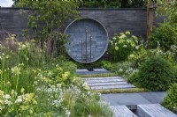 Un endroit pour se retrouver. Hampton Court Flower Festival 2021. Une cour urbaine est plantée dans des tons reposants de gris, vert et blanc. Une fontaine circulaire sur le mur recycle les tuyaux et les robinets.