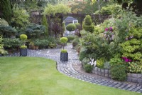 Vue sur jardin avec arbres matures, conifères, plantes vivaces et annuelles dans les parterres de fleurs d'été avec pelouse et patio décoratif fait de galets récupérés
