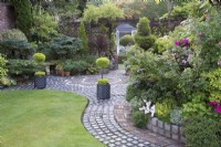 Vue sur jardin avec arbres matures, conifères, plantes vivaces et annuelles dans les parterres de fleurs d'été avec pelouse et patio décoratif fait de galets récupérés