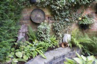 Mur de briques et parterre de fleurs surélevés en matériaux récupérés et décorés d'anciens équipements de jardinage et de jardinières et plantés d'Hedera, de fougères et d'Hosta
