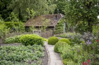 Un chemin pavé simple bordé de briques mène à travers un jardin de style cottage avec des légumes et des fleurs de chaque côté