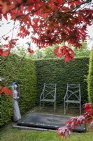 Couverture d'if entoure Sculpture d'ange en bois d'if par John Aulman dans un jardin secret. Deux élégantes chaises en métal sont formellement disposées à côté. Acer palmatum 'Atropurpureum' au premier plan