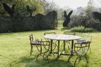 Table et chaises sur la pelouse de Balmoral Cottage, Kent en avril, encadrées par des oiseaux topiaires émergeant du haut des haies environnantes de buis et d'ifs.