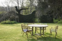 Table et chaises sur la pelouse de Balmoral Cottage, Kent en avril, encadrées par des oiseaux topiaires émergeant du haut des haies environnantes de buis et d'ifs, et une glycine standard.