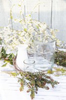 Grands et petits pots en verre, eau, plumes, saule pleureur, bâtons de bouleau, brindilles couvertes de lichen, brins d'hébé et bâtons de prunellier en fleurs disposés sur une table