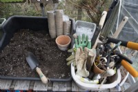 Une table de rempotage dans une serre se trouve à côté d'une cuve contenant divers outils de jardinage, notamment un maillet, des cisailles, des ciseaux et plusieurs paires de gants de jardinage. Un paquet de pots écologiques est posé sur la table de rempotage.