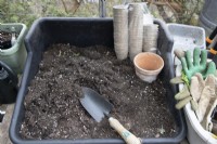 Une table de rempotage dans une serre est prête avec du compost, des pots de plantes écologiques et une truelle. Les gants de jardinage sont rangés sur le côté.
