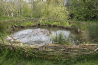 Un étang dans un cadre naturel avec une clôture de platebande extérieure basse faite de branches torsadées et de matériaux en bois.