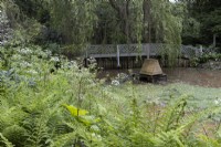 Vue sur un étang avec des plantations au bord de l'eau et une maison de canard en bois au milieu de l'étang. Lewis Cottage, jardin NGS Devon. Le printemps.