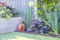 Heuchera 'Ginger Peach' plantée dans un parterre de fleurs à côté d'un ornement de poisson en métal rustique